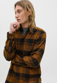 Młoda osoba z długimi blond włosami w brązowej i czarnej koszuli w kratę pozuje z ręką przy brodzie na jednolitym tle.