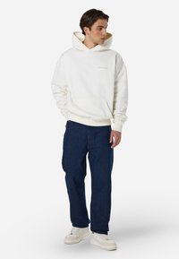 Λευκή φούτερ με κουκούλα και μπροστινές τσέπες, με ανοιχτό μπεζ κείμενο, συνδυασμένη με σκούρα μπλε τζιν και λευκά sneakers. Χαλαρή εφαρμογή και σχέδιο.