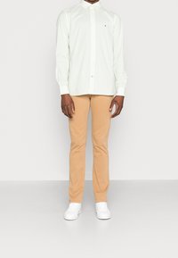 Camicia button-up verde chiaro con colletto, abbinata a pantaloni slim-fit beige e sneakers bianche. La camicia presenta un motivo sottile e un dettaglio del logo.