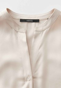 Blusa beige a maniche corte con scollatura rotonda, chiusura con bottoni e texture liscia. L'etichetta riporta "un giorno." Design pulito e semplice.