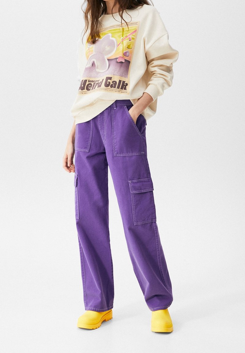 Femme portant un sweat-shirt graphique beige, un pantalon cargo violet et des bottes jaune vif, debout avec une main dans la poche.
