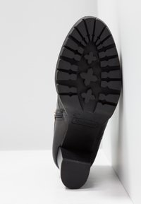 Sort støvlesål med dybt slidbanemønster, der inkluderer tre blomsterformede greb og en blokhæl mod en hvid hjørnet baggrund.