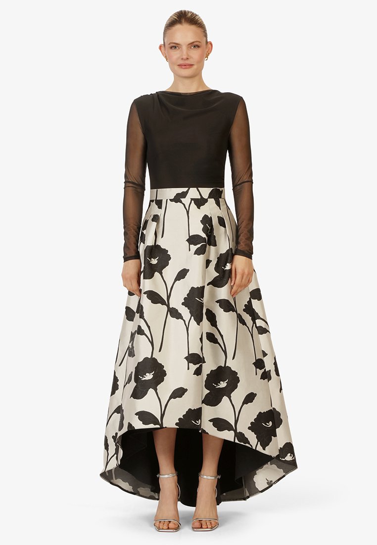 Zwart langemouwen top met doorschijnende mouwen, gecombineerd met een high-low rok met een crème achtergrond en een zwart bloemenpatroon. Zilveren haksandalen.