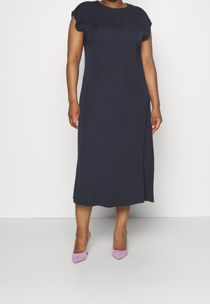 Kvinde iført en enkel, kortærmet, navyblå midikjole kombineret med lavendelfarvede spidse hæle på en ensfarvet hvid baggrund.