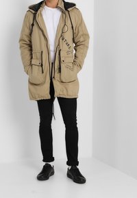 Khaki parka z kapturem ze sznurkiem, dwoma przednimi kieszeniami, ozdobnym czarnym napisem oraz czarną podszewką, w połączeniu z czarnymi spodniami i trampkami.