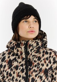 Beige gesteppte Jacke mit schwarzen und braunen abstrakten Flecken, ausgestattet mit einem hohen Kragen, einem Reißverschluss und verstellbaren Manschetten. Schwarze Strickmütze.