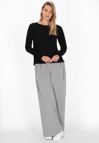 Top negru cu maneci lungi și guler rotund, asociat cu pantaloni gri cu picioare largi. Modelul poartă adidași albi și stă pe un fundal simplu.