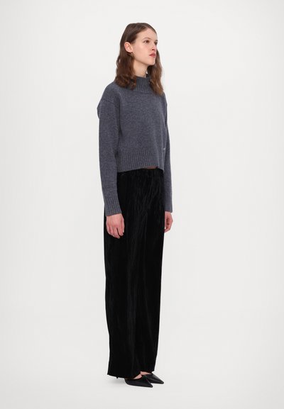 Pull en gris coupé avec un col côtelé, associé à un large pantalon en velours noir. Le modèle se tient de côté, mettant en valeur la texture du tissu et l'ajustement.