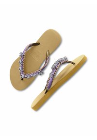 Des tongs avec une semelle en caoutchouc beige, des brides tressées violettes ornées d'ornements scintillants et une semelle intérieure texturée pour une meilleure adhérence.