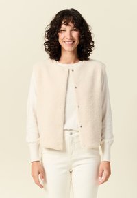 Crème fleece vest met drukknoopjes, ronde halslijn en zonder mouwen; gedragen over een lichte trui, gecombineerd met crème broeken.