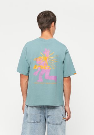 Kraft t-skjorte i teal med et dristig flerfarget grafisk trykk på baksiden, som har elementer i gult, rosa og oransje. Løs passform.