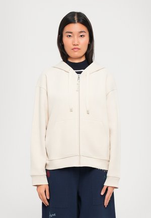 Giovane donna con capelli neri lisci che indossa una felpa cremosa con zip e pantaloni blu navy con testo colorato, in piedi contro uno sfondo chiaro e uniforme.