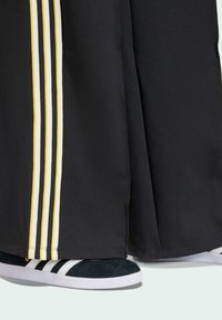 Svarta träningsbyxor med tre vertikala ränder i vitt och gult, kombinerat med svarta mockaskor med vita ränder och vit sula.