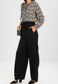 Pantaloni beige a vita alta e a gamba larga abbinati a una blusa a fantasia floreale bianca e nera con un collo con fiocco. Accessoriati con una borsa verde.