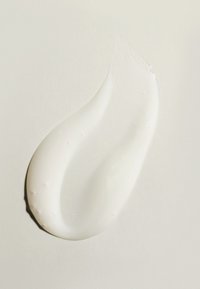 Weiße Creme oder Gel in einer glatten, fließenden Form mit glänzender Oberfläche und kleinen Luftblasen auf einem hellbeigen strukturierten Hintergrund.