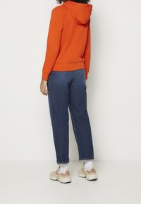 Pomarańczowa bluza z kapturem o luźnym kroju, zestawiona z niebieskimi dżinsami. Stylizacja uzupełniona beżowo-różowymi sportowymi butami i białymi skarpetkami.