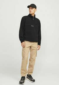 Black fleece pullover with a quarter zip and logo detail on the chest, paired with beige cargo pants and black sneakers.