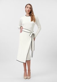 Knit dress in off-white with black trim, featuring a wrap design and tied belt accent at the waist. Long sleeves and fitted silhouette.