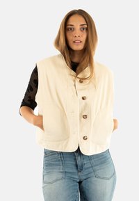 Beige corduroy vest met een ronde kraag, knopen aan de voorkant en twee grote zakken. Draag het over een zwart langarmshirt en combineer het met blauwe jeans.
