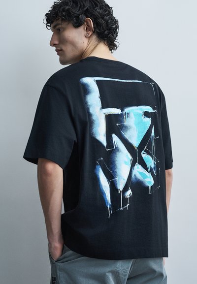 Giovane uomo con capelli ricci scuri che indossa una t-shirt nera oversize con una grafica astratta blu e bianca sulla schiena, abbinata a pantaloni grigi.