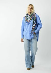 Chemise boutonnée bleu clair associée à un jean délav�é. Le modèle porte un foulard coloré avec des motifs géométriques et floraux.