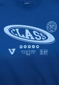 Modré bavlněné tričko s bílým potiskem textu, včetně "CLASS" a "DENIM", s logem a rokem založení "EST 97" výrazně zobrazeným.
