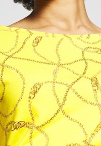 Gul topp med ett kedjemönster i guldtoner. Har en vid halsringning och en strukturerad yta som betonar designen av sammanlänkade länkar.