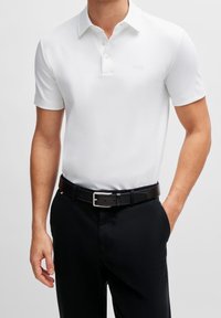 Polo blanc à manches courtes avec col, comportant trois boutons et un logo subtil sur la poitrine. Ceinture noire et pantalon foncé.