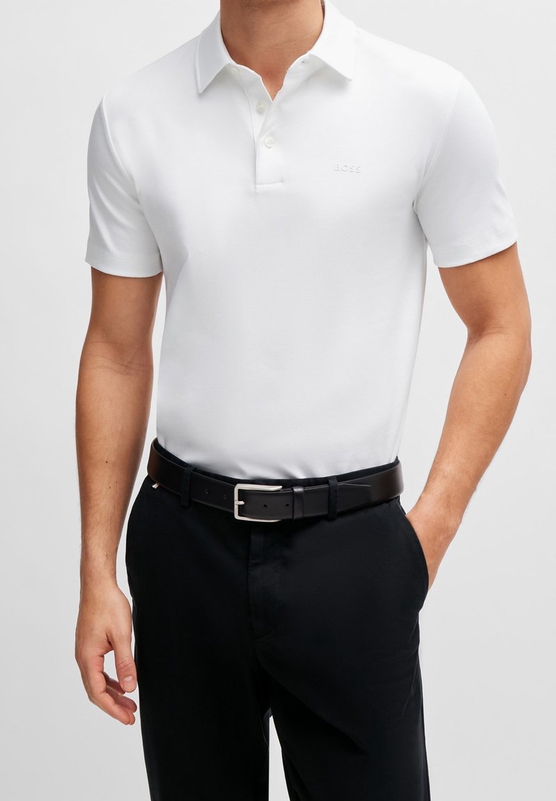 Polo blanc à manches courtes avec col, comportant trois boutons et un logo subtil sur la poitrine. Ceinture noire et pantalon foncé.