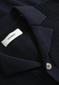 Sötét tengerészkék kötött kardigán gombos záródással, bordázott gallérral és belül egy fehér címkével, melyen a "GABBA" felirat olvasható.