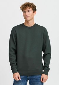 Mørkegrønn sweatshirt i rund halsdesign, med lange ermer og ribbestrikkede mansjetter, laget av et mykt, teksturert stoff. Bært med denimjeans.
