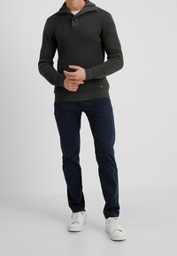 Suéter de punto verde oscuro con un cuello alto y detalle de botones, combinado con pantalones marinos y zapatillas blancas, de pie frente a un fondo blanco.