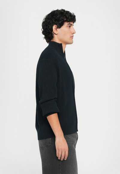 Mörk marinblå räfflad zip-up tröja med hög krage, visad på en manlig modell i profil, över grå jeans.