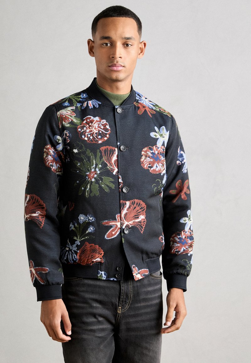 Λουλουδάτο bomber jacket με μαύρο φόντο, έντονα πολυχρωμα λουλούδια, ριμπ μανσέτες και κουμπωτή κλείσιμο. Συνδυασμένο με σκούρες τζιν.