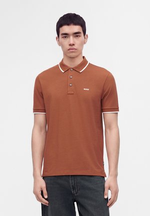 Ung mand iført en rustfarvet polo-shirt med hvide kanter og tre knapper, stående mod en ensfarvet lys baggrund.