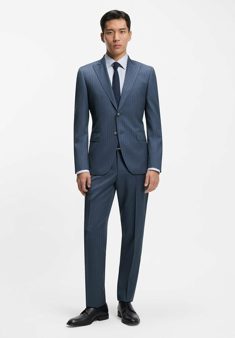 Granatowy garnitur w prążki wykonany z gładkiej tkaniny, dopasowany fason z dwoma guzikami, klapy z nacięciem i spodnie z prostymi nogawkami; zestawiony z jasną koszulą w paski.