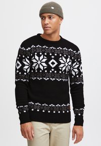 Black knit sweater featuring a white and gray snowflake pattern, ribbed cuffs and hem, worn with a gray beanie. Cream-colored pants.