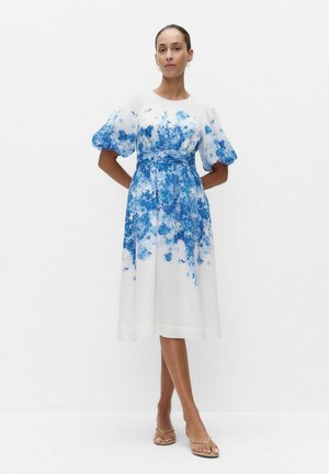 Kvinde stående med hænderne bag ryggen iført hvid midi-kjole med blåt blomsterprint og pufærmer, beige sandaler.