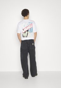 Weißes T-Shirt mit einem auffälligen grafischen Druck in Pink, Blau und Schwarz auf der Rückseite, kombiniert mit schwarzen, weiten Cargo-Hosen.