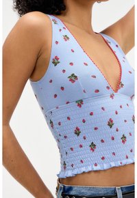 Ljusblått ribbat crop top med djup V-ringning, prydd med små jordgubbsmönster och en volangkant. Röda stygn detaljerar.