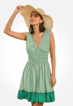 Mujer con un vestido verde sin mangas y estampado, con un gran sombrero de paja, sujetando el borde del sombrero con los ojos cerrados, fondo neutro.