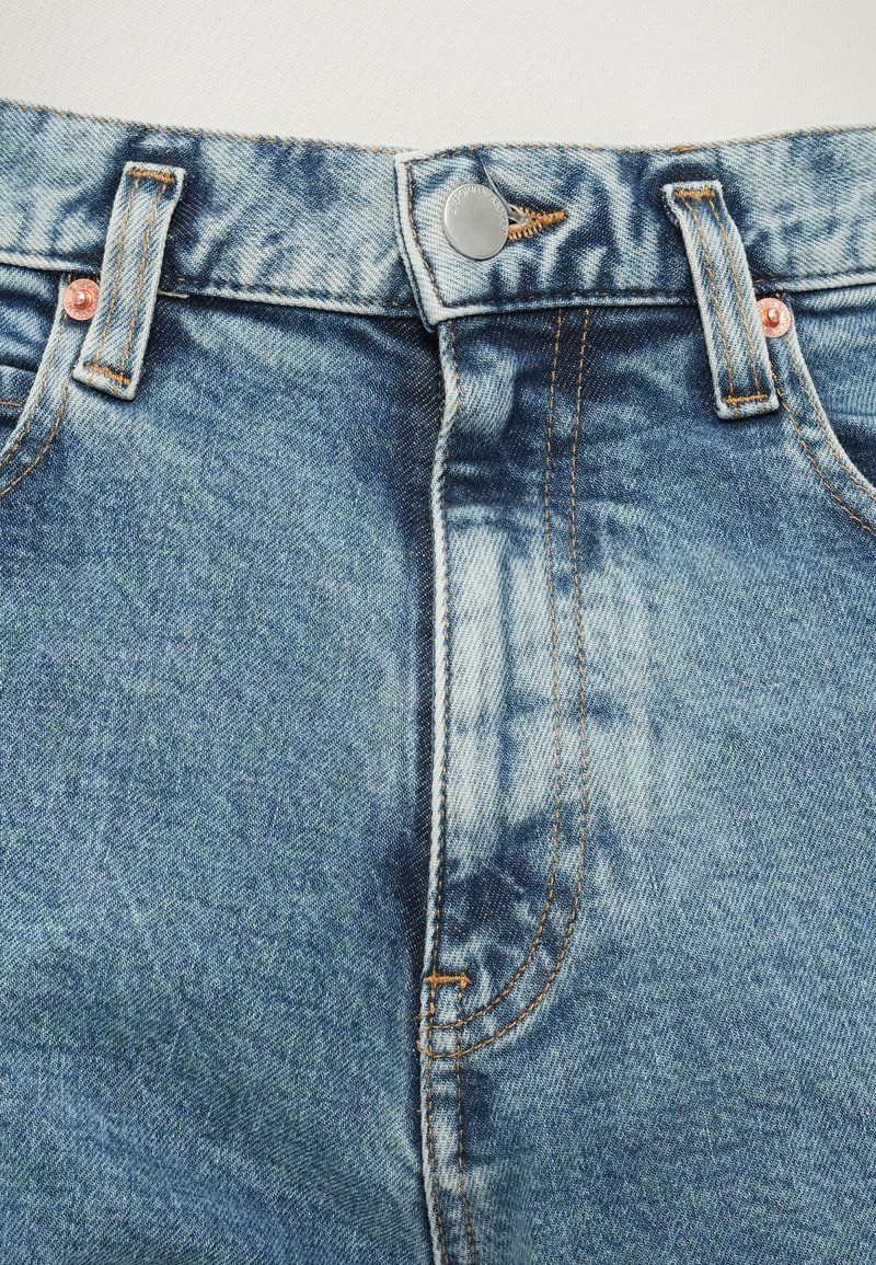 Medium wash denim jeans with copper rivets, five-pocket design, orange stitching, and a button closure at the waist.