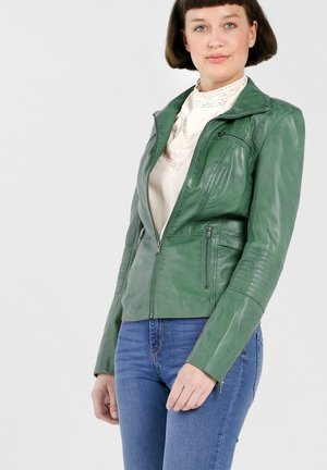 Vrouw die een groene leren jas draagt over een witte kanten blouse en blauwe jeans, staand tegen een effen witte achtergrond.