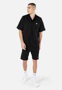 Maglietta nera a maniche corte con tessuto testurizzato, chiusura con bottoni e piccolo logo sul petto, abbinata a pantaloni corti neri e sneakers bianche.