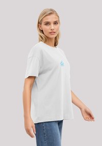 Weißes Baumwoll-T-Shirt mit kurzen Ärmeln, das ein hellblaues grafisches Design auf der Brust zeigt. Kombiniert mit blauen Jeans.