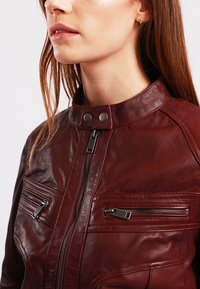 Burgundy leather jacket with a high collar, zip closure, and two zippered chest pockets. Features smooth texture and metallic hardware accents.