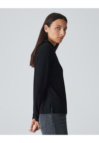 Schwarzes, strukturiertes Longsleeve-Shirt mit taillierter Passform und seitlichen Schlitzen. Kombiniert mit grauen Jeans, ausgestattet mit einem Rundhalsausschnitt und dezentem Detail.
