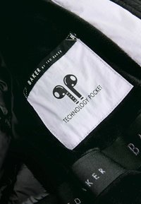 Crna tkanina enterijera s bijelom etiketom na kojoj piše "BAKER by TED BAKER" i "TEHNOLOŠKA DŽEP", ukrašena dizajnom slušalica.