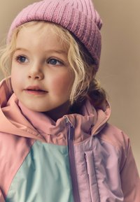 Mångfärgad vinterjacka med rosa, lila och mintavsnitt; har en dragkedja framtill och en huva. Rosa stickad mössa på modellens huvud.
