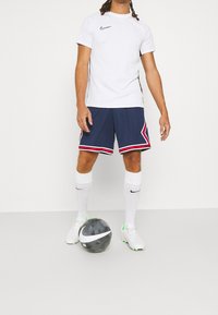 Vit kortärmad Nike-tröja, marinblå shorts med röda och vita ränder, vita strumpor och vita fotbollsskor med gröna detaljer. Svart och vit fotboll.
